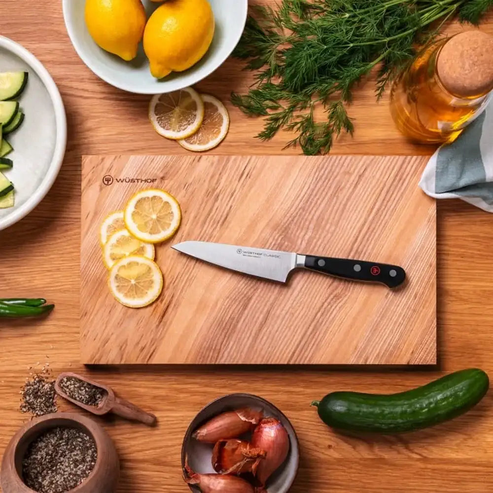 Wooden cutting board with a Wusthof knife, surrounded by lemons, herbs, and vegetables on a wooden surface.