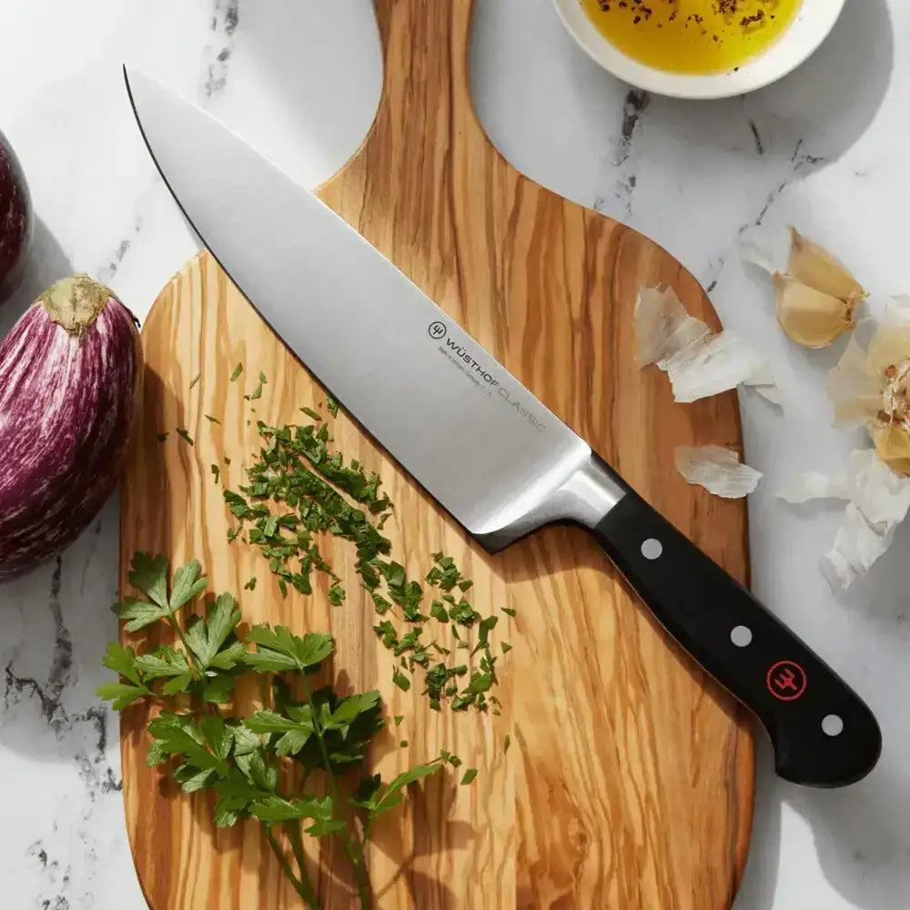 Wooden cutting board with a chef's knife, chopped herbs, and a small bowl of oil on a marble surface.