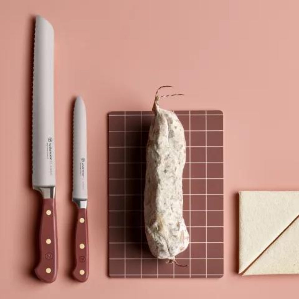 Two  knives with red handles, a block of cheese on a grid board, and a white envelope on a pink background.