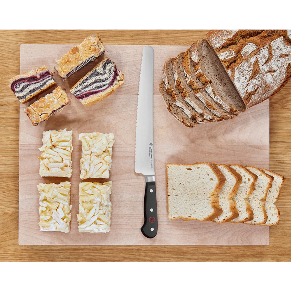 A serrated bread knife with a black handle is displayed on a light wooden cutting board, surrounded by sliced bread, cheese cubes, and fruit bars on a wooden table.