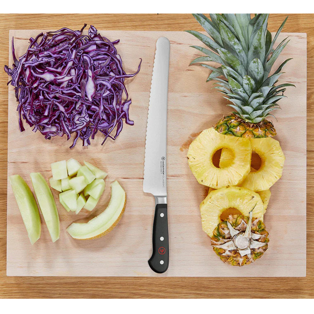 A serrated stainless steel kitchen knife with a black handle lies on a light wood cutting board, surrounded by food props including shredded purple cabbage, sliced green apple, and halved pineapple rings.