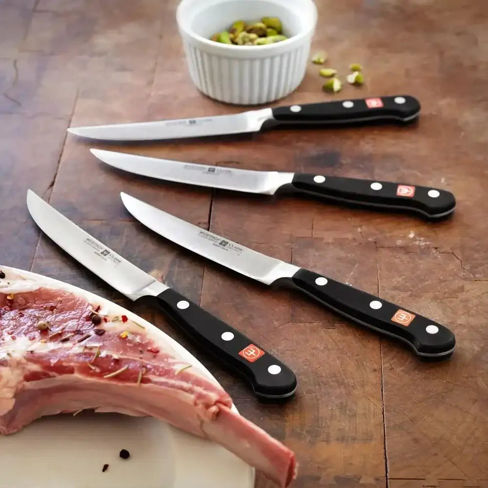 Set of knives with black handles on a wooden surface next to a piece of meat and a small bowl.