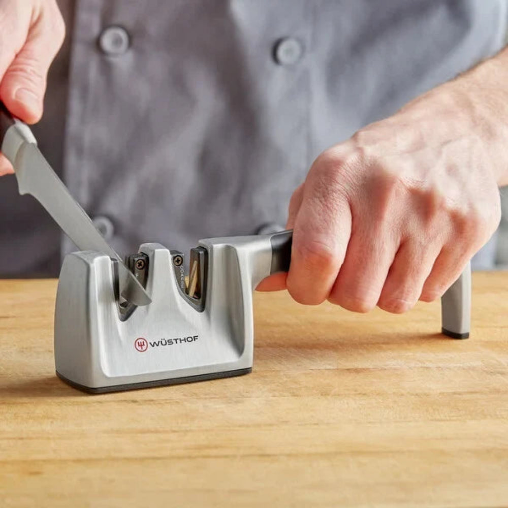 A close-up of a person using a Wüsthof knife sharpener on a wooden cutting board, with the person's hands and gray chef coat visible.