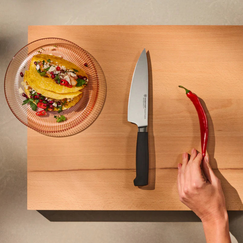 Wooden cutting board with a plate of tacos, knife, and red chili pepper on a neutral background