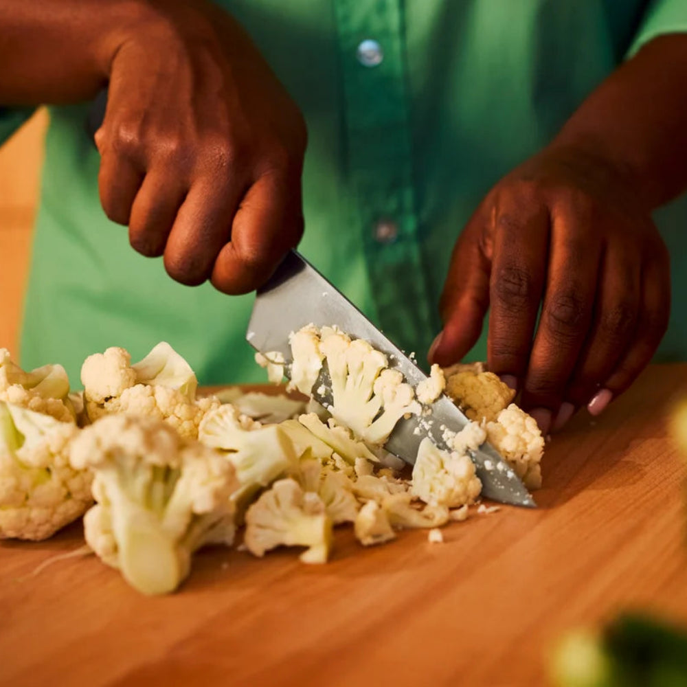 Person cutting cauliflower on a wooden cutting board with a knife.