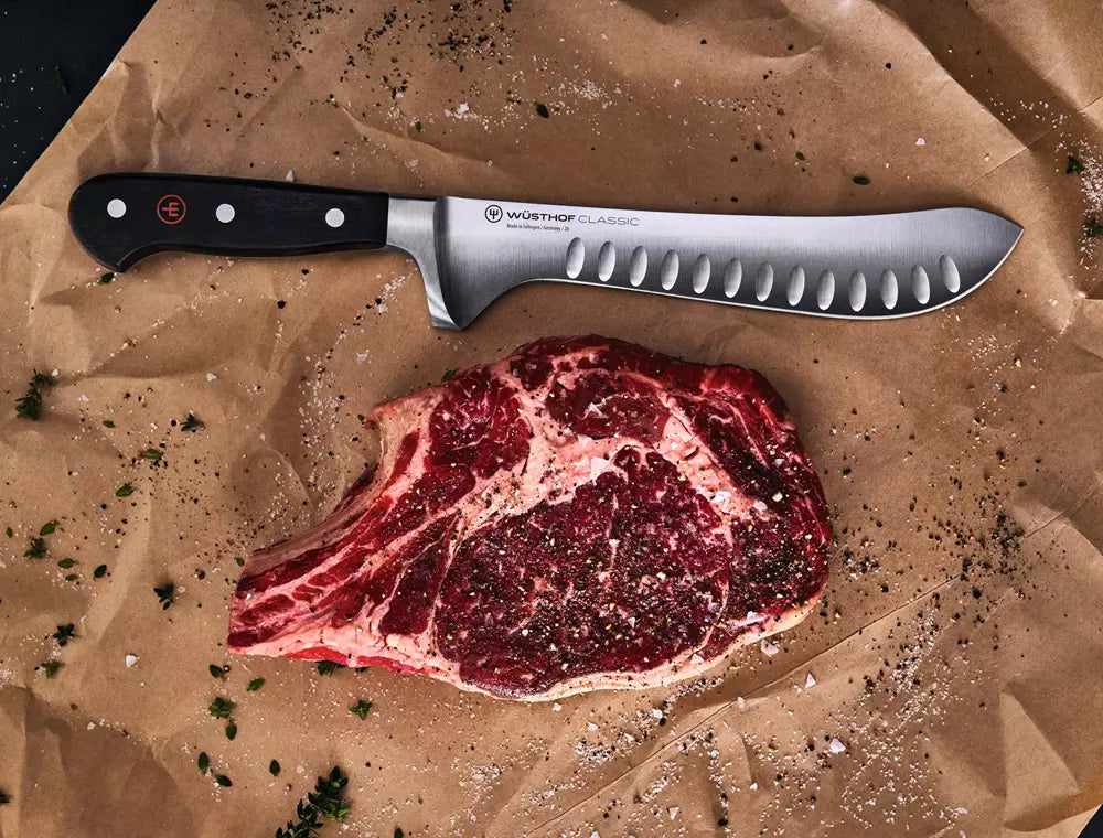 A large Workhof butcher knife with a black handle and serrated stainless steel blade, laid diagonally on brown butcher paper next to a raw marbled steak seasoned with herbs and salt.