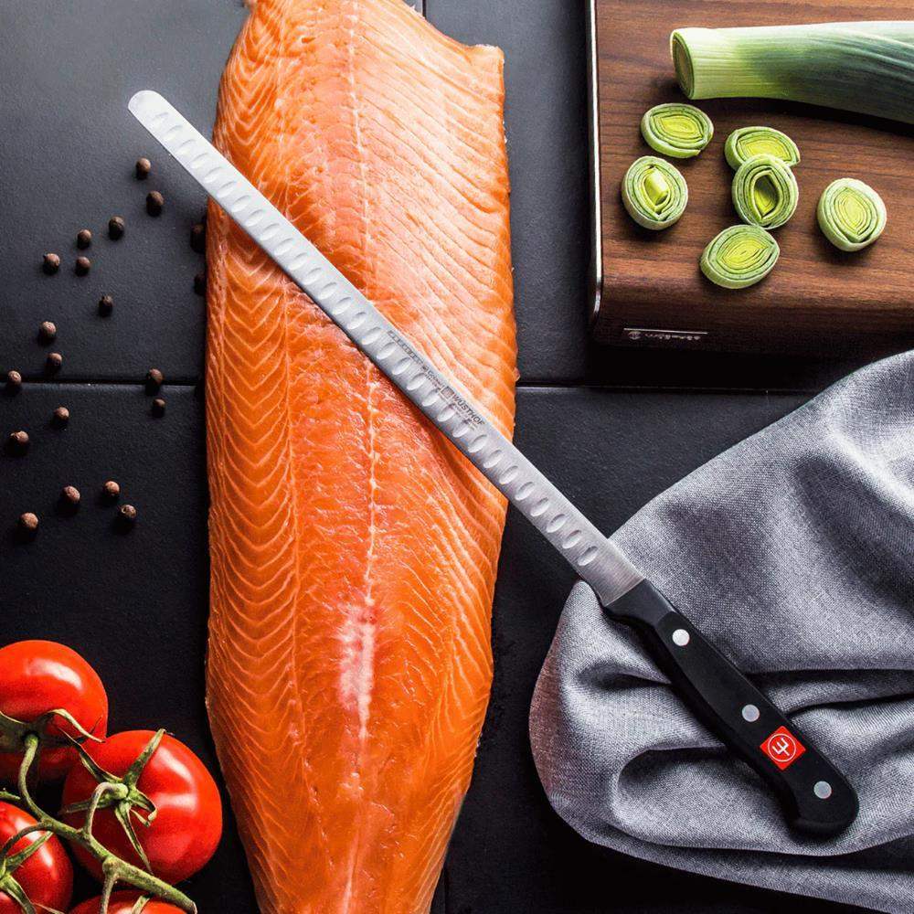 Raw salmon fillet with a knife, leeks, tomatoes, and a gray towel on a dark surface.