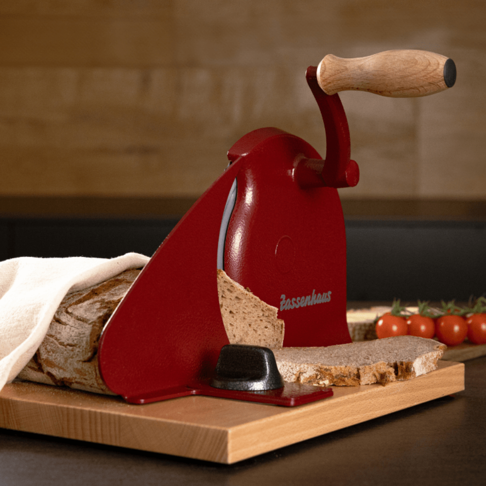 A red manual bread slicer with a wooden handle, shown slicing a loaf of bread on a wooden board with sliced bread and tomatoes nearby.
