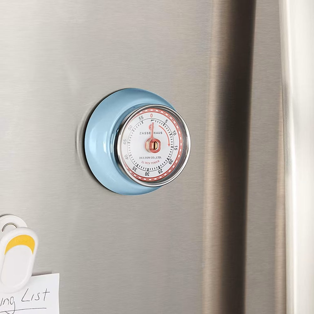 A round kitchen thermometer with a light blue rim and a white dial face, mounted on a brushed stainless steel refrigerator door next to a white clip and sticky note.