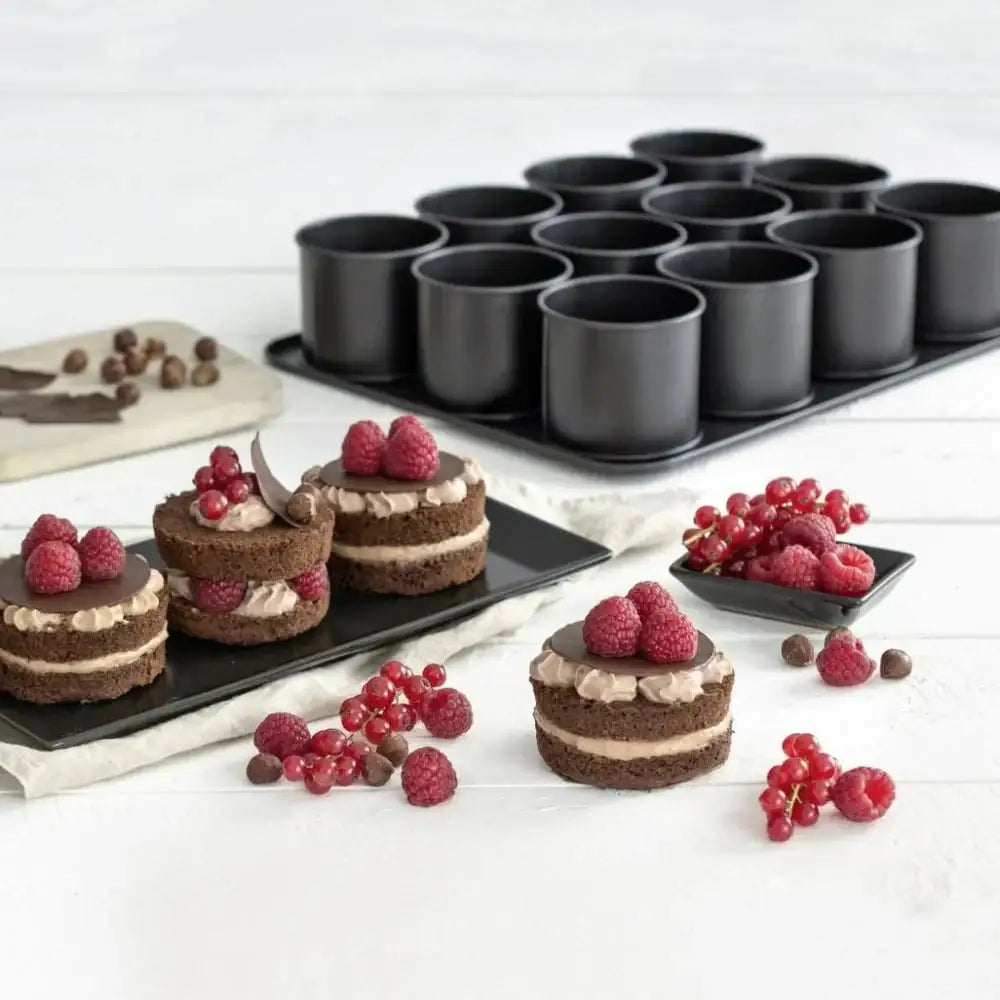 Small chocolate cakes with raspberries on a white surface, surrounded by black cake pans.