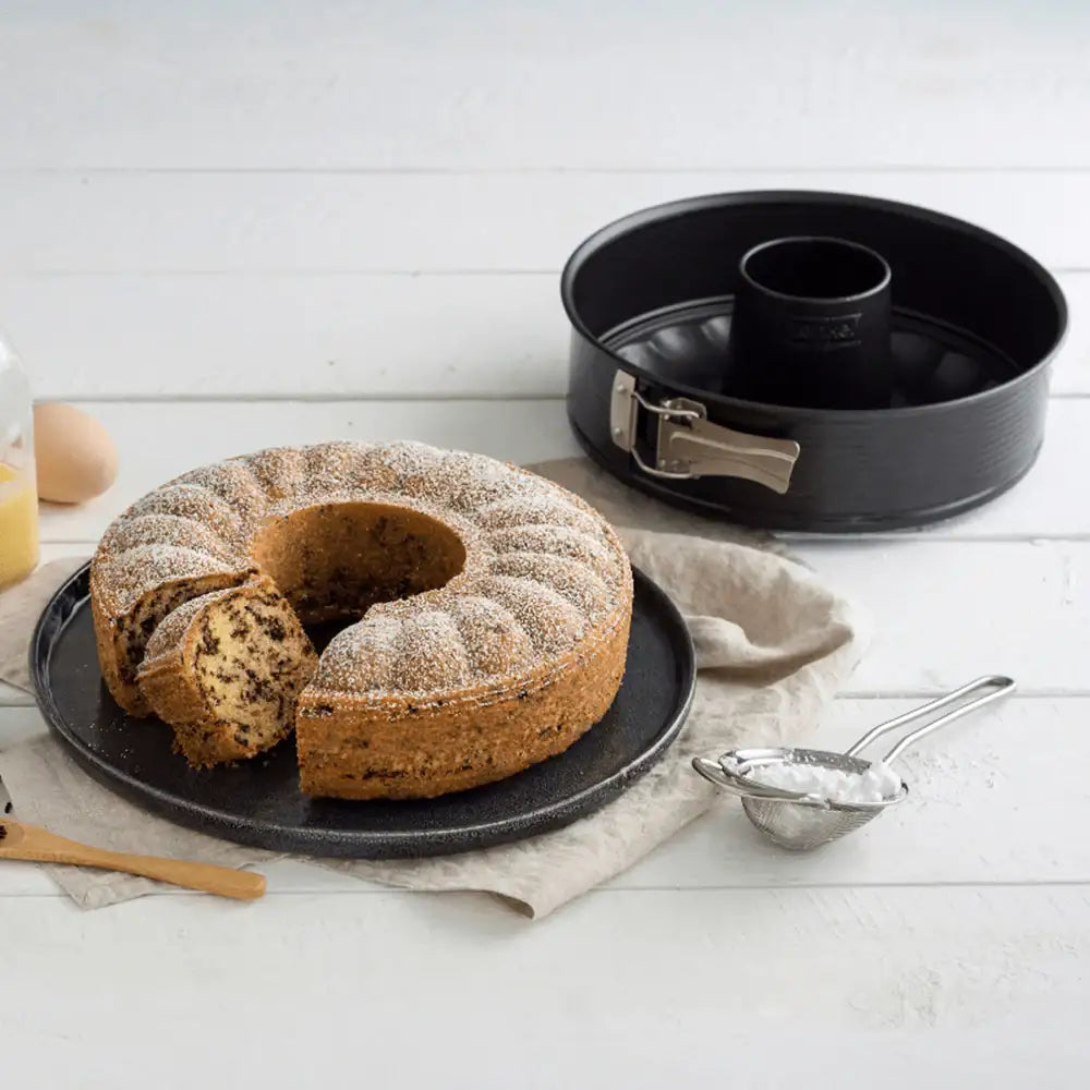 Bundt cake on a plate with a baking pan and sifter in the background on a white surface.