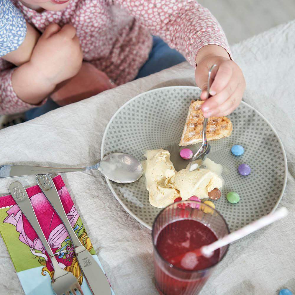 Child eating dessert with a fork and spoon, surrounded by colorful candies and a glass of juice on a textured surface.