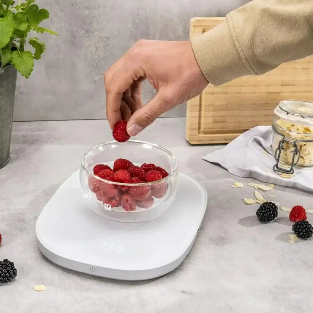 Person adding raspberries to a glass bowl on a white surface with a neutral background