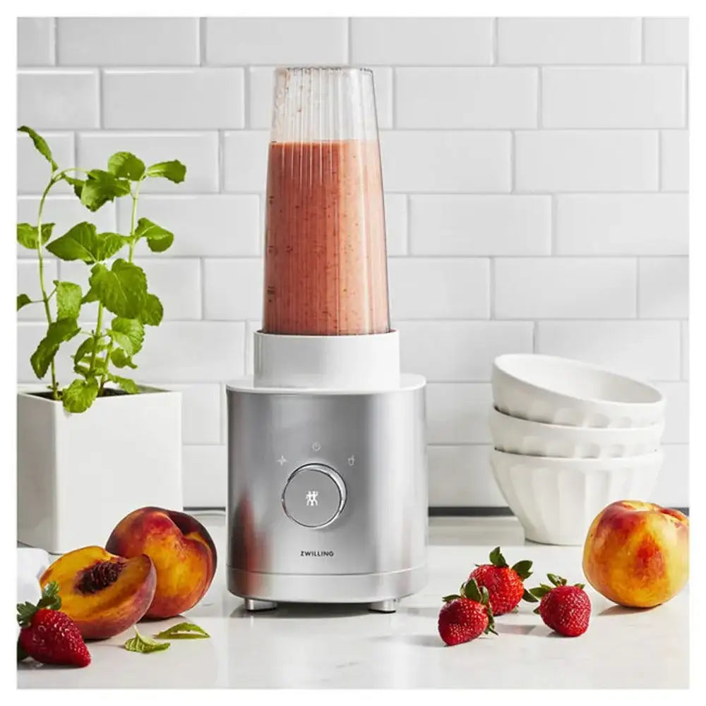 Blender with a smoothie surrounded by fruits and bowls on a kitchen counter.