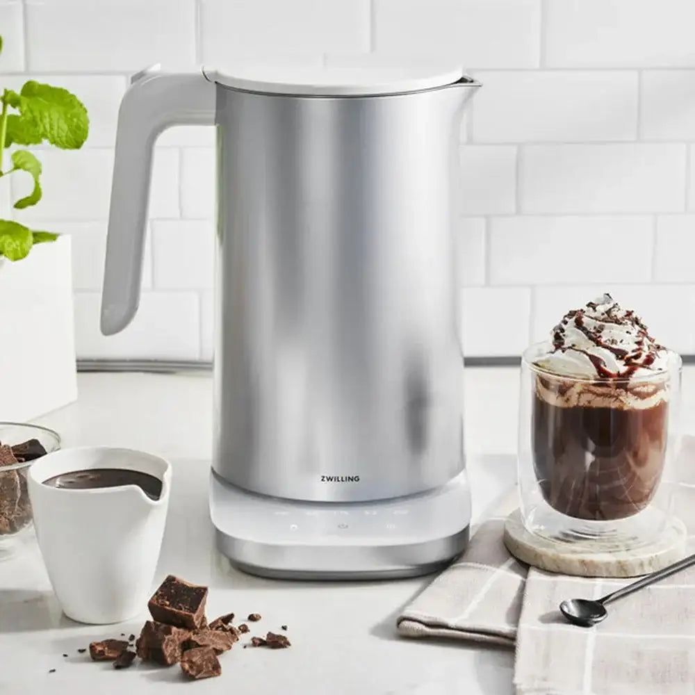 Silver electric kettle on a kitchen counter with a glass of hot chocolate and chocolate pieces.