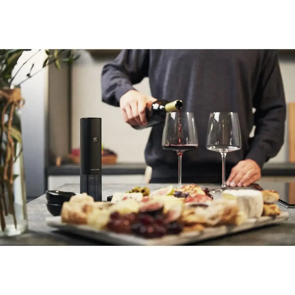 Person pouring wine into two glasses with a bottle of wine and food on a table.