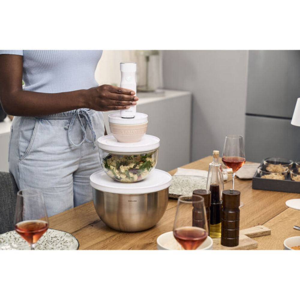 Person using a kitchen appliance on a dining table with glasses and bottles in the background