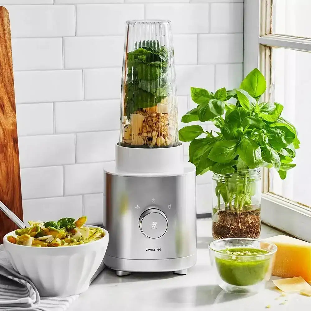 Blender with green ingredients on a kitchen counter, surrounded by a bowl of salad, a plant, and a glass of green juice.