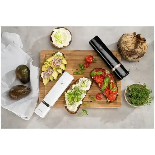 Wooden cutting board with slices of bread topped with avocado, tomato, and other ingredients, along with a knife and vegetables on a light gray surface.