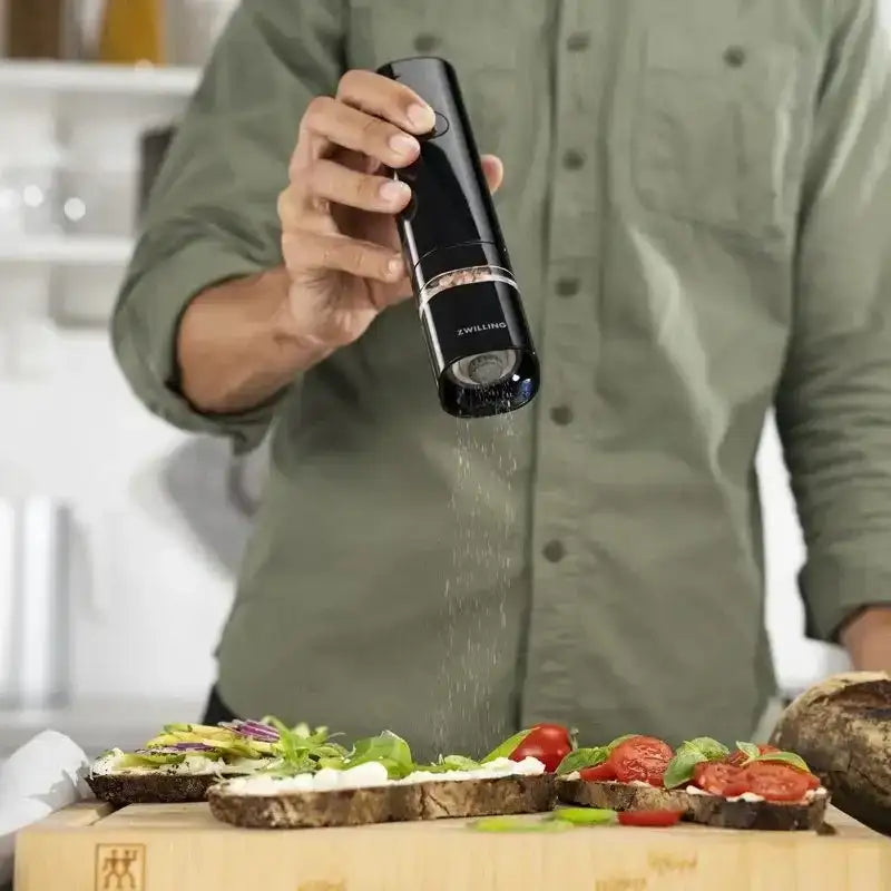 Person seasoning a salad with a pepper grinder in a kitchen setting