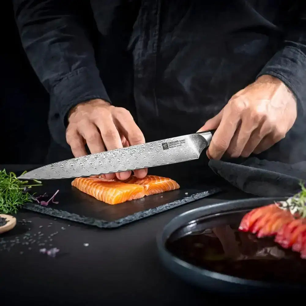 Person using a chef's knife to cut salmon on a dark surface with a bowl of sauce nearby.