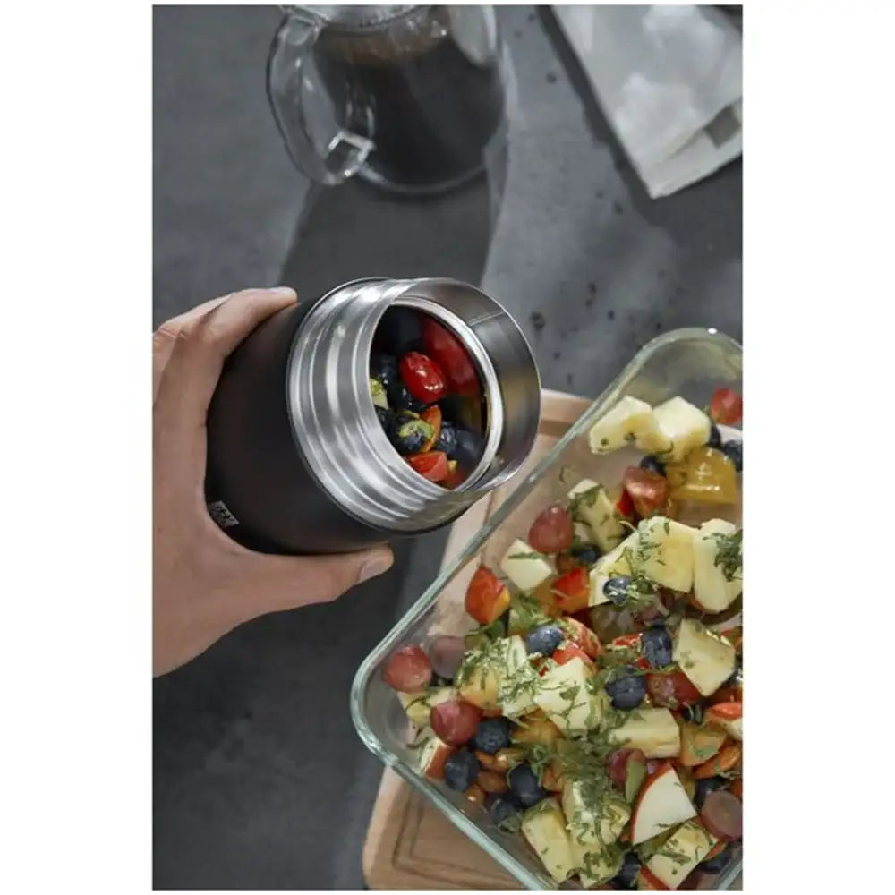 Hand holding a insulated food container with salad next to a glass dish of salad on a gray surface.