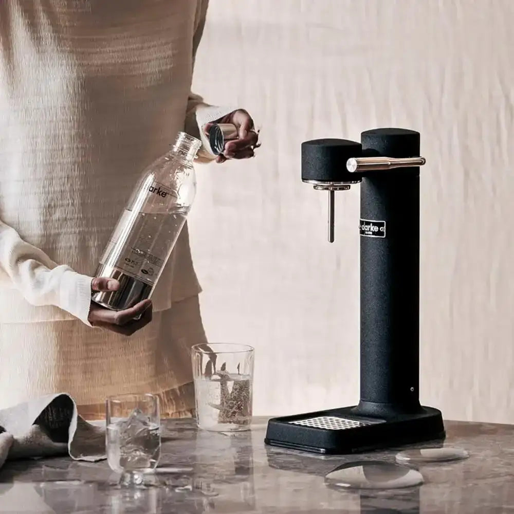 Aarke III Premium Water Carbonator in black on kitchen counter, person holding bottle ready for carbonation, modern home setting from Cookin Stores.