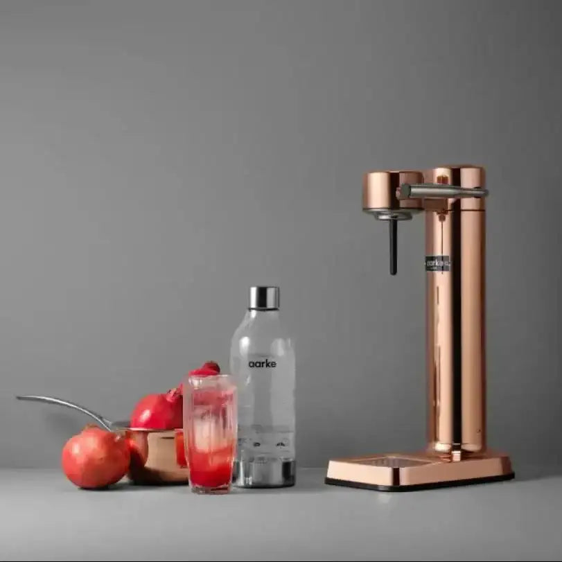 Aarke III premium copper water carbonator with glass, bottle, and pomegranates on grey background