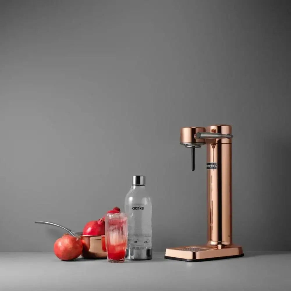 Aarke III premium copper water carbonator with glass, bottle, and pomegranates on grey background