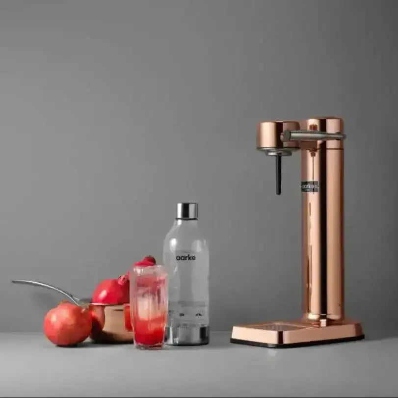 Copper water carbonator with bottle, glass of sparkling drink, and pomegranates on gray background