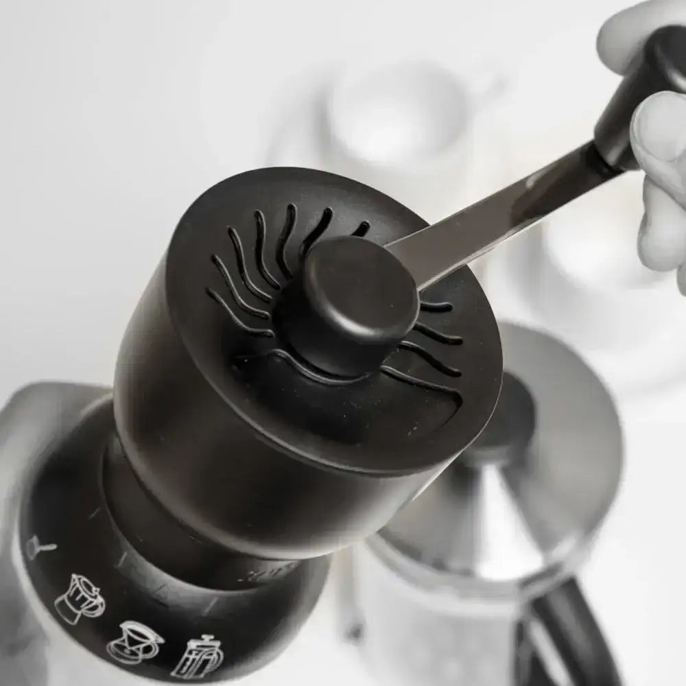 Black coffee grinder with a hand holding a spoon, against a blurred background