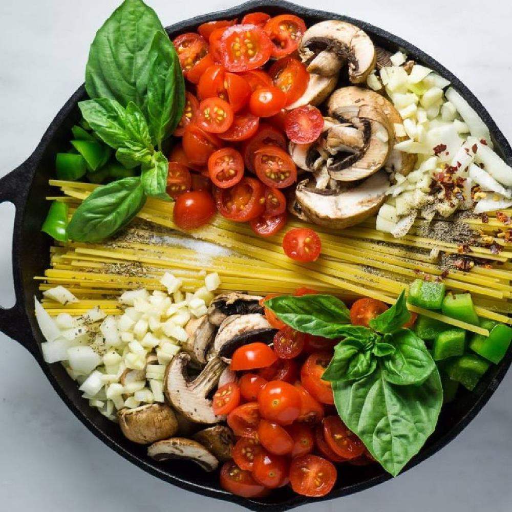 Amt 38cm non-stick braising pan with uncooked pasta, cherry tomatoes, mushrooms, basil, peppers, onions and spices, on a marble surface for Cookin Stores kitchenware.