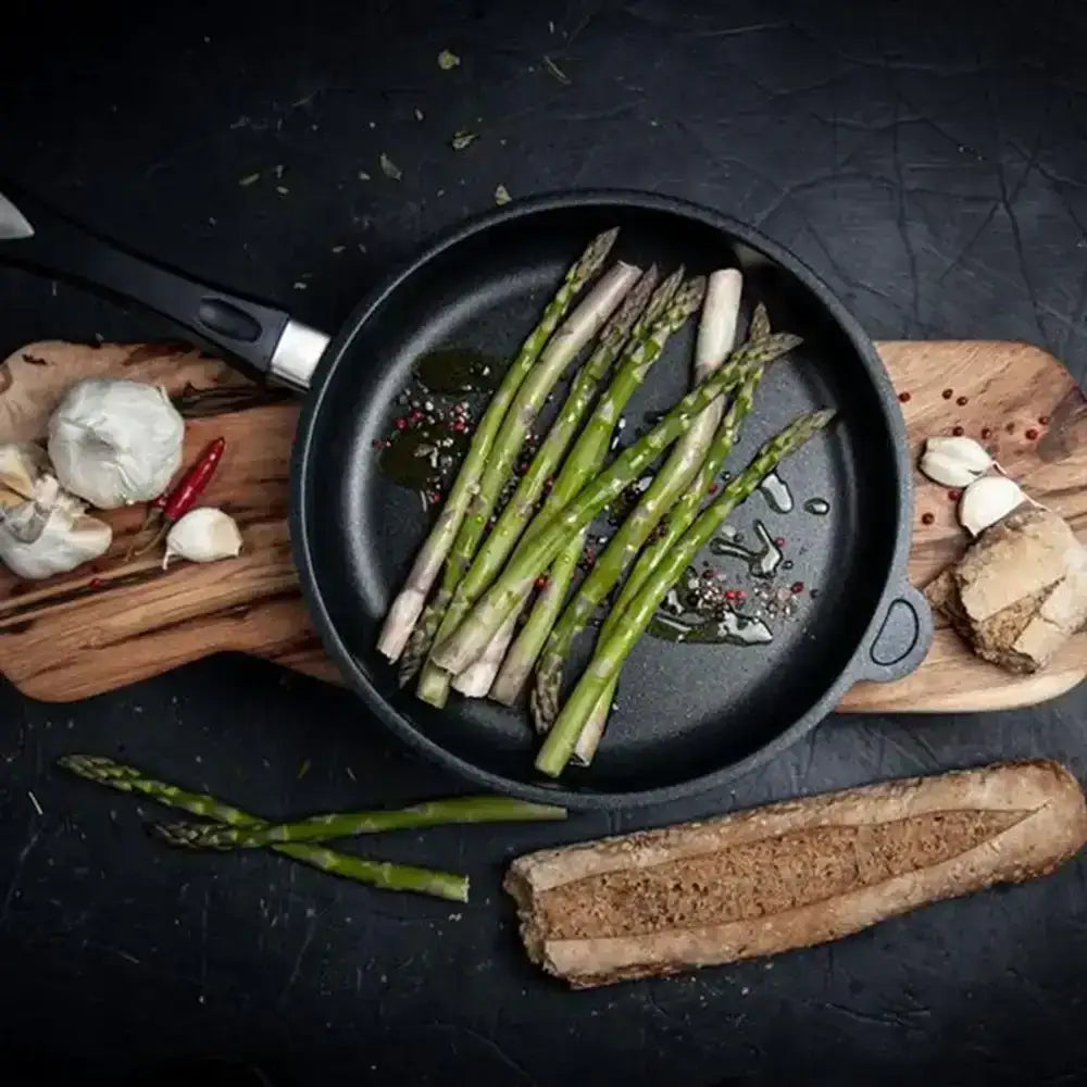 Asparagus in a frying pan on a dark surface with bread and garlic.