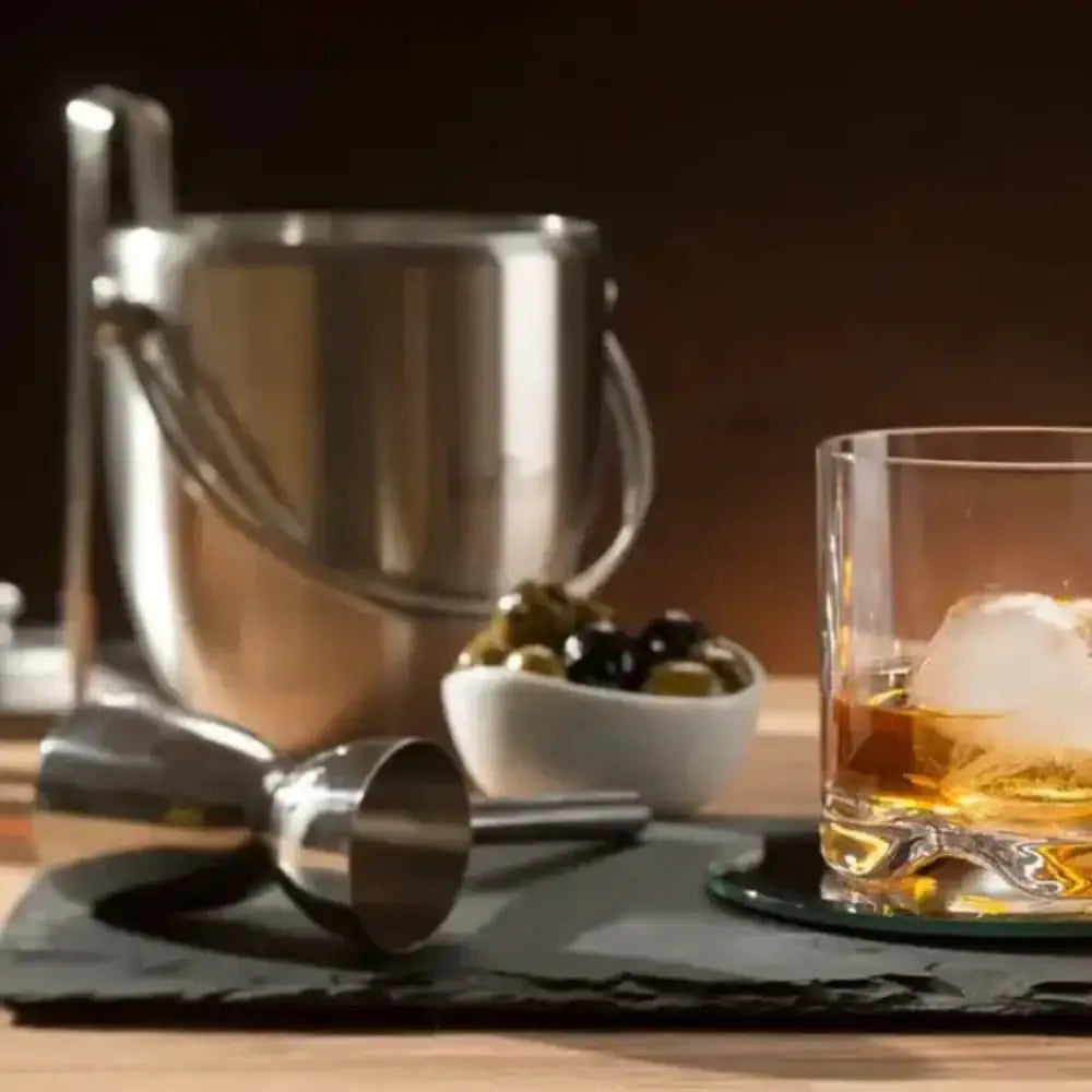 Glass of whiskey with ice on a wooden table, accompanied by a cocktail shaker and bowl of olives.