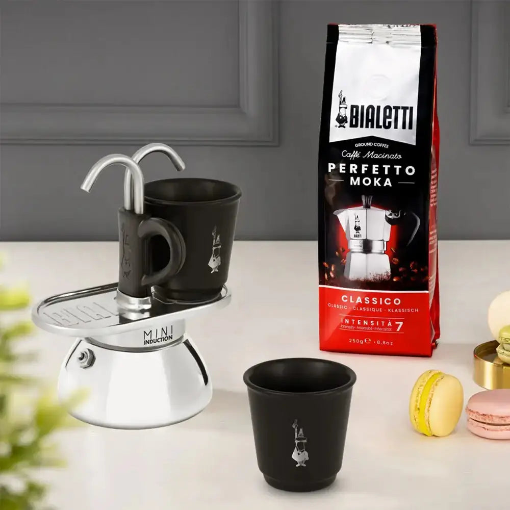 Bialetti coffee maker with two black cups and a box of Bialetti coffee on a light surface.