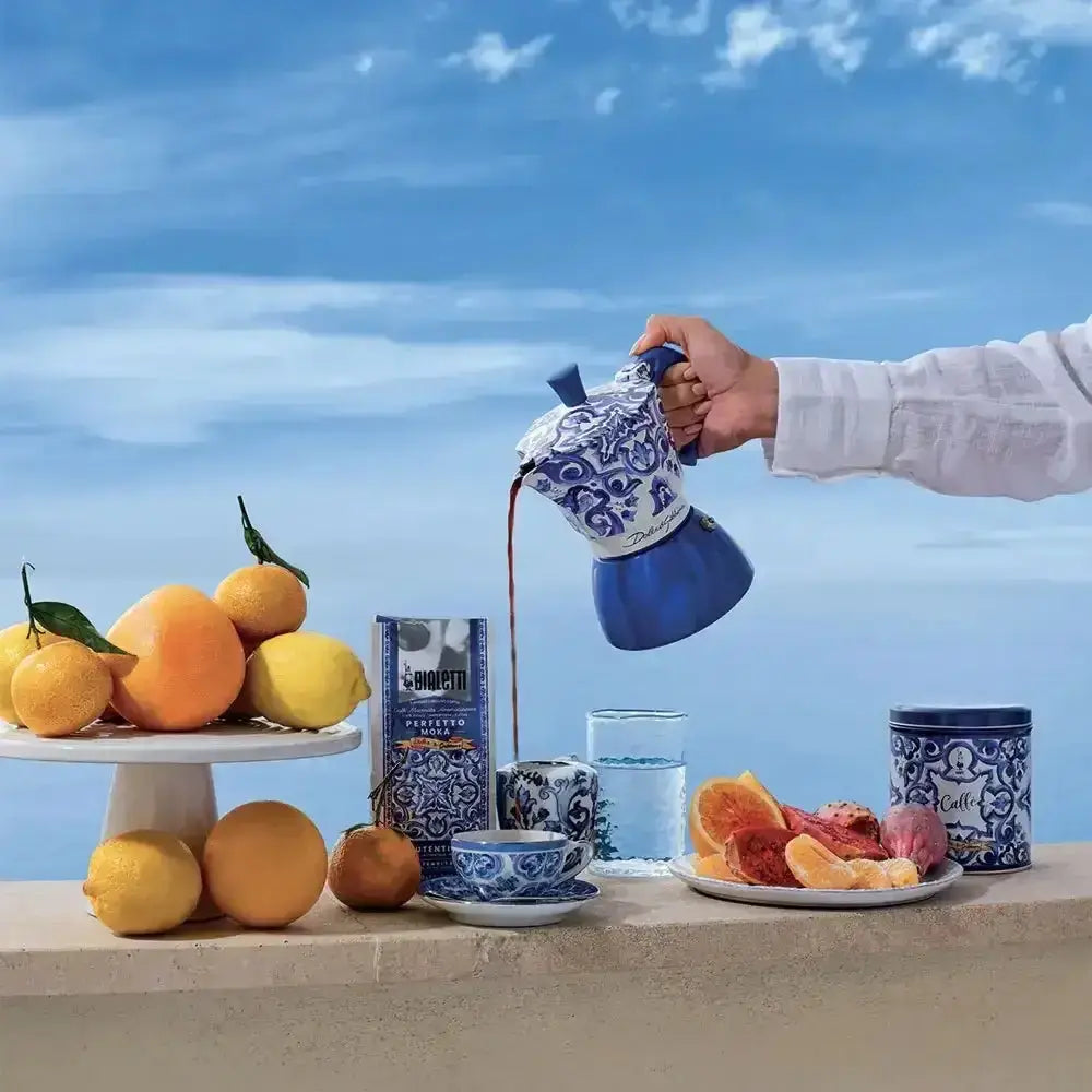 Bialetti Dolce & Gabbana Mediterraneo moka pot pouring coffee by lemons, oranges, and blue-patterned cups outdoors