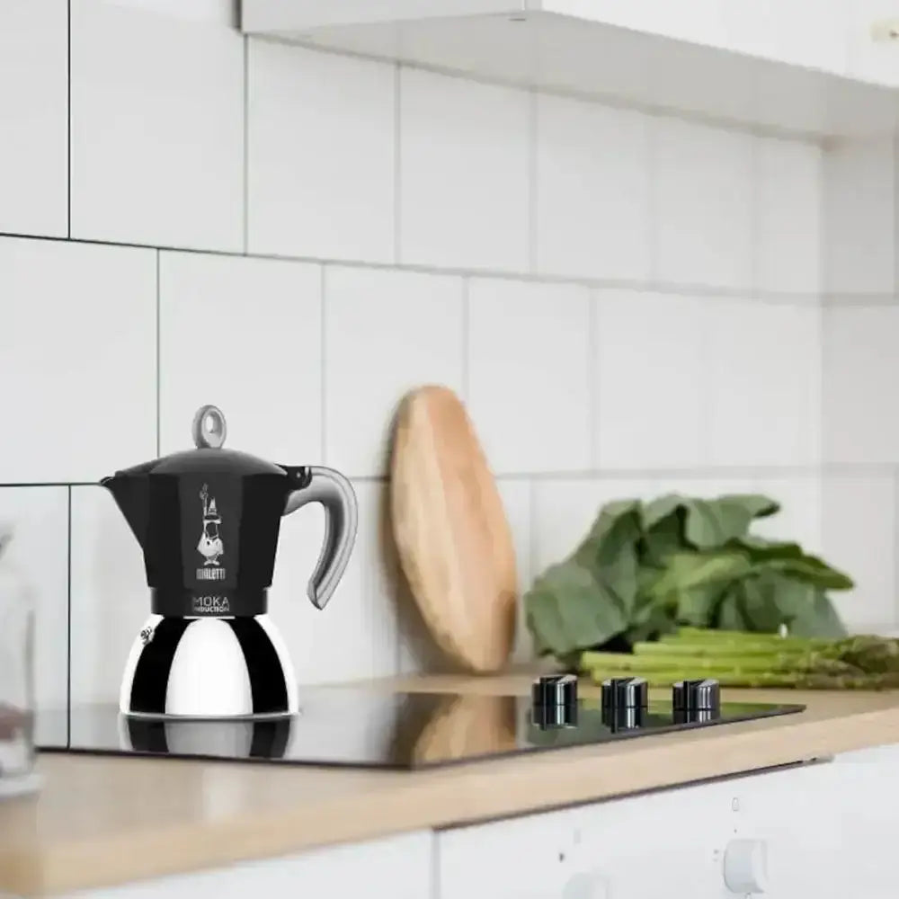 Black and silver espresso maker on a kitchen counter with a tiled wall and green vegetables in the background.