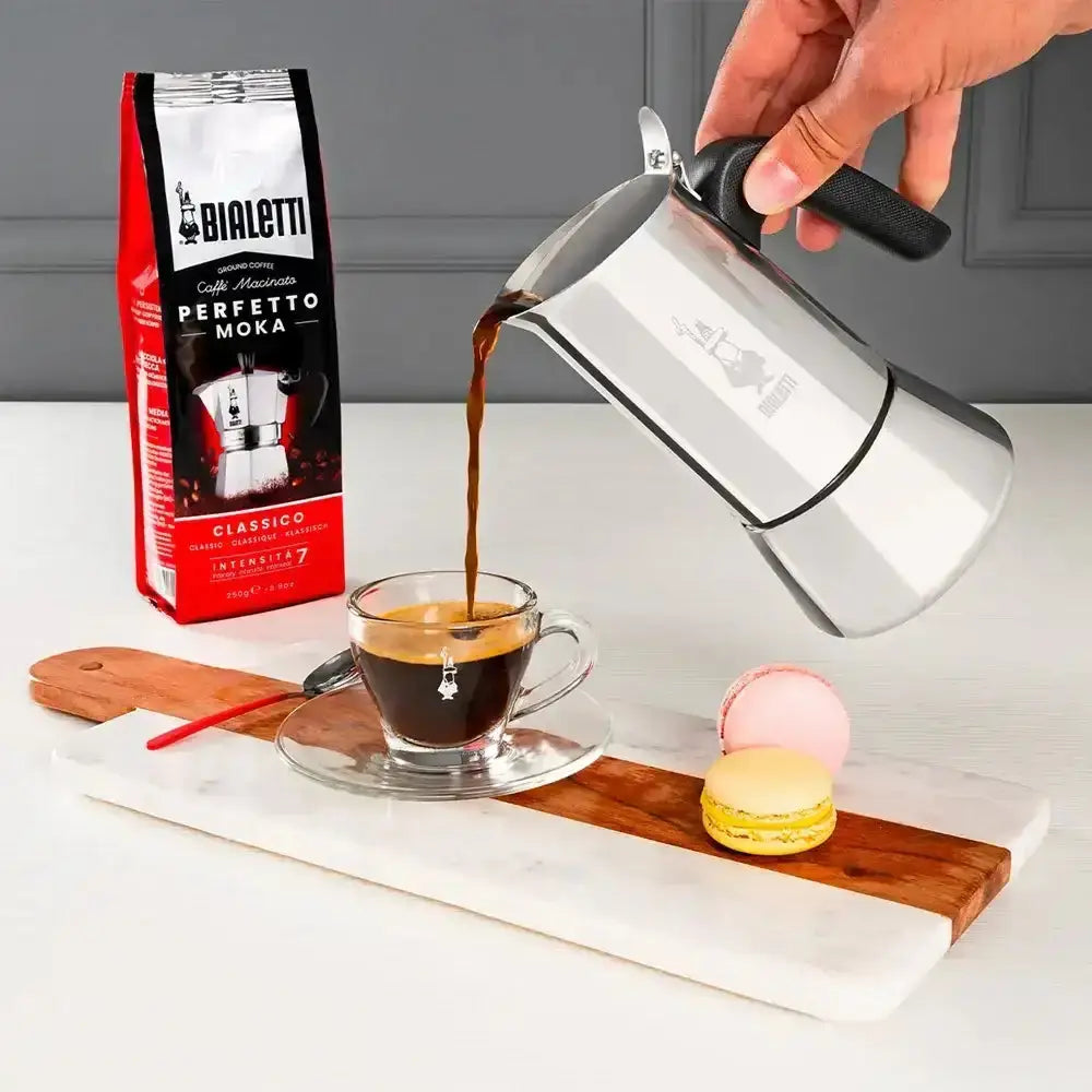 Bialetti coffee maker being used to pour coffee into a cup with a Bialetti coffee bag and macarons in the background.