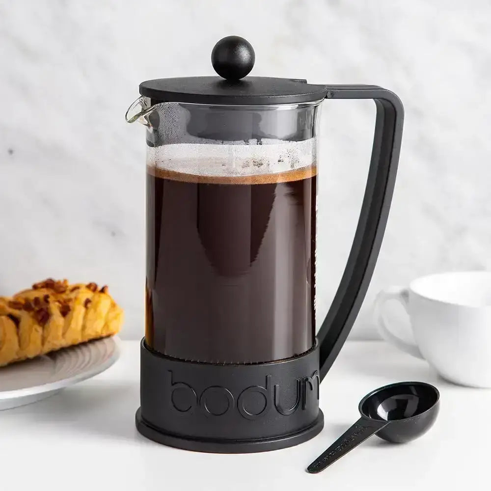 Black Bodum French press with coffee on a white surface with pastries and a cup in the background.