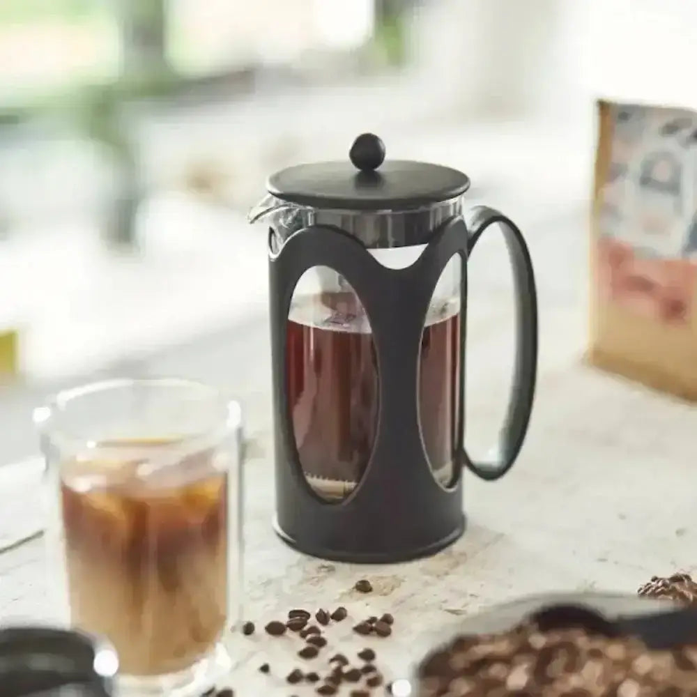Black French press with a glass of coffee on a table with coffee beans.