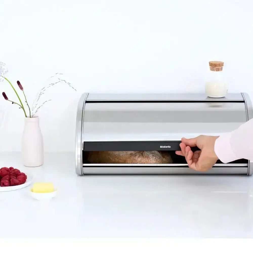 Stainless steel bread box on a white surface with a hand opening it, surrounded by berries and a vase.