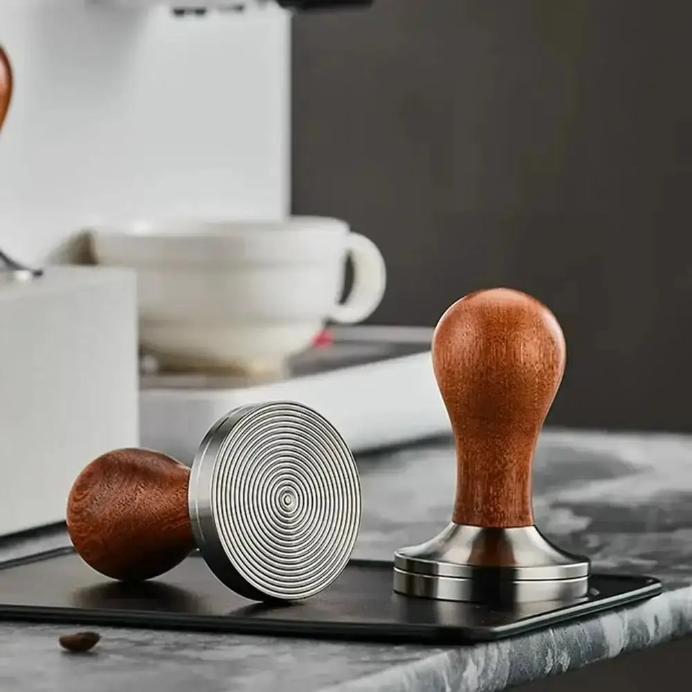 Two wooden coffee tampers on a marble surface with a blurred coffee-making setup in the background.