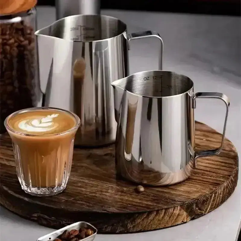 Two stainless steel milk pitchers on a wooden tray with a glass of coffee and coffee beans.
