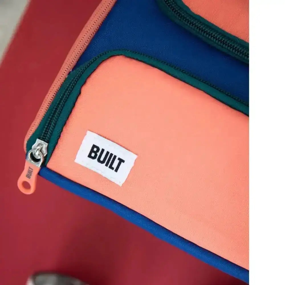 A vibrant coral and navy blue insulated lunch bag with green zipper trim and built brand label on front