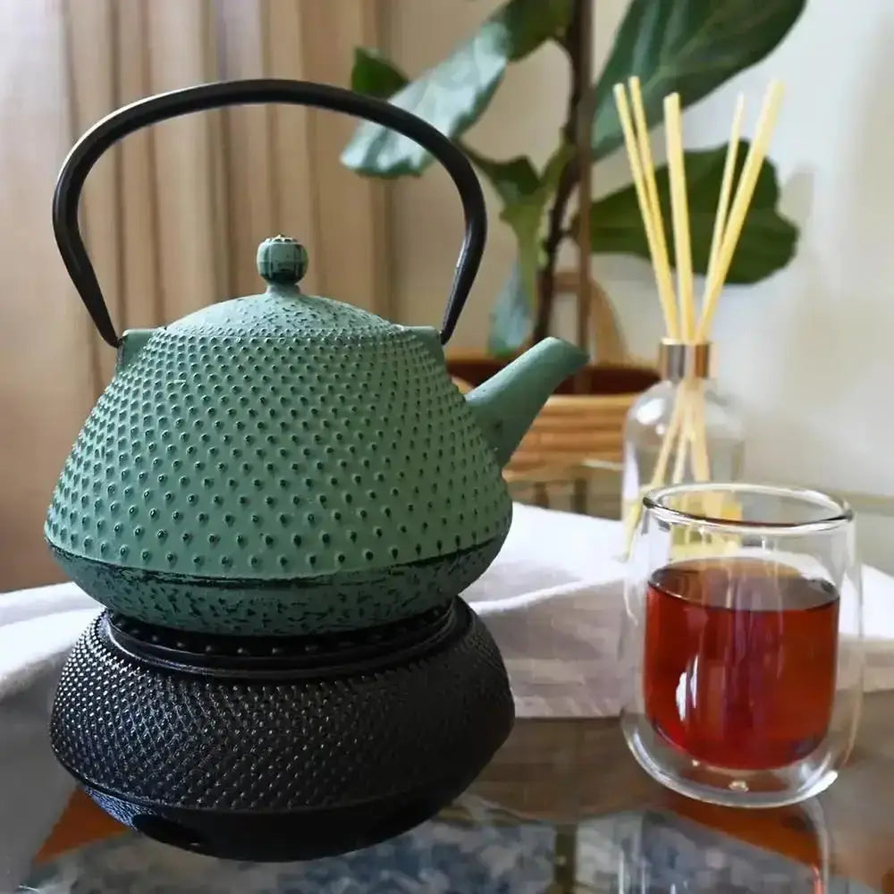 Green teapot on a black stand with a glass of tea in the background