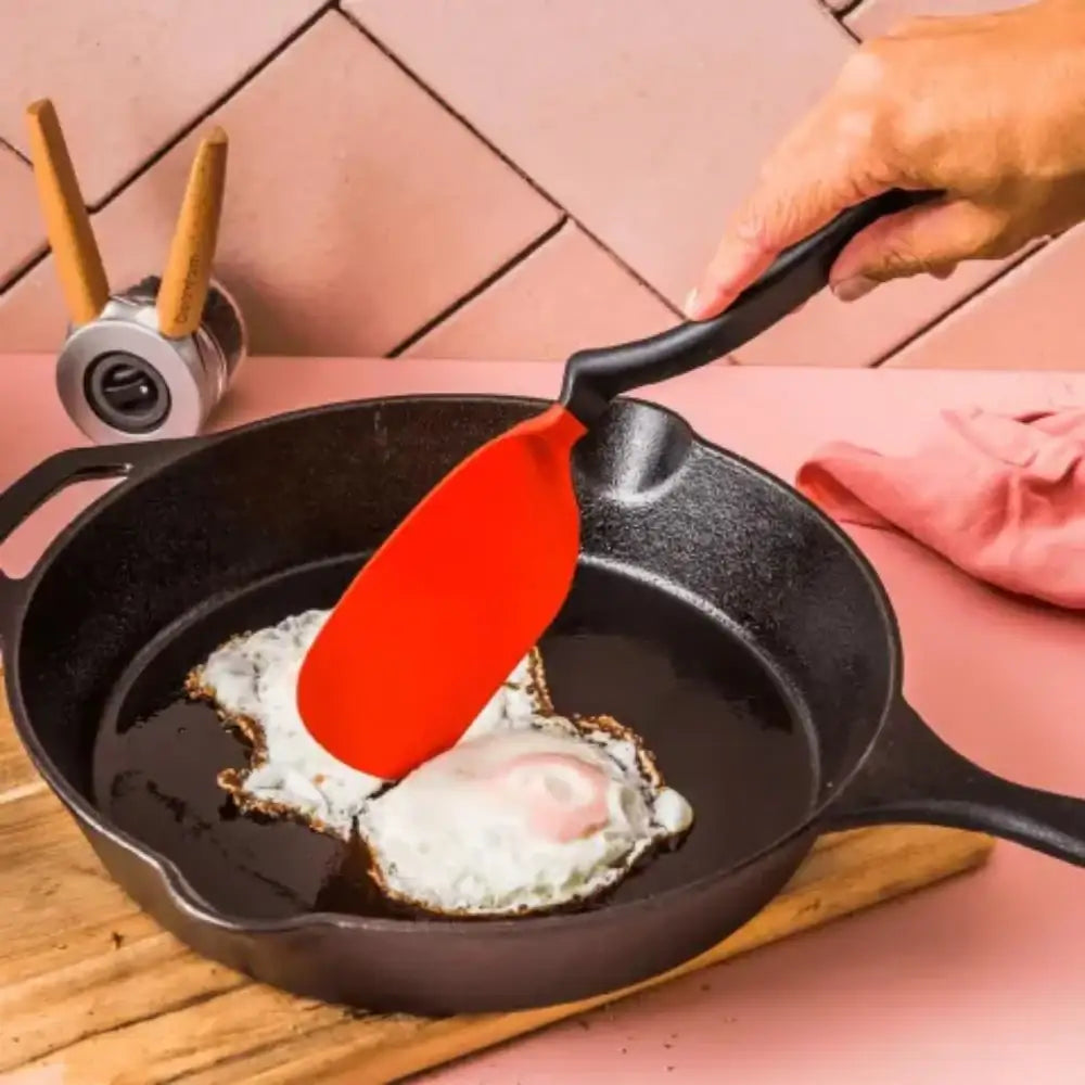 Person frying eggs in a cast iron skillet with a red spatula, on a pink tiled surface.