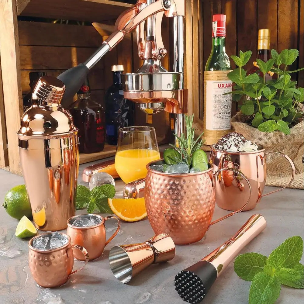 Copper cocktail shaker set with bottles, glasses, and ingredients on a wooden shelf.