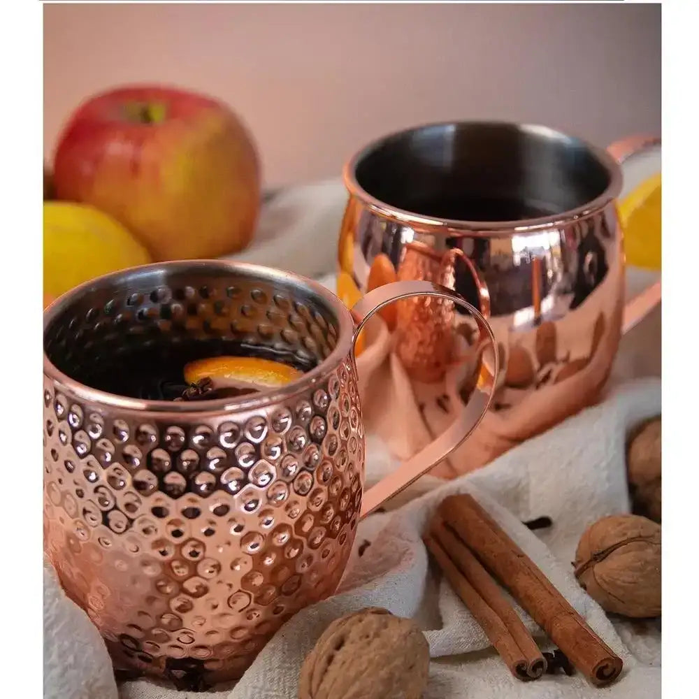 Two copper mugs with a warm beverage, surrounded by apples, oranges, cinnamon sticks, and walnuts on a soft surface.