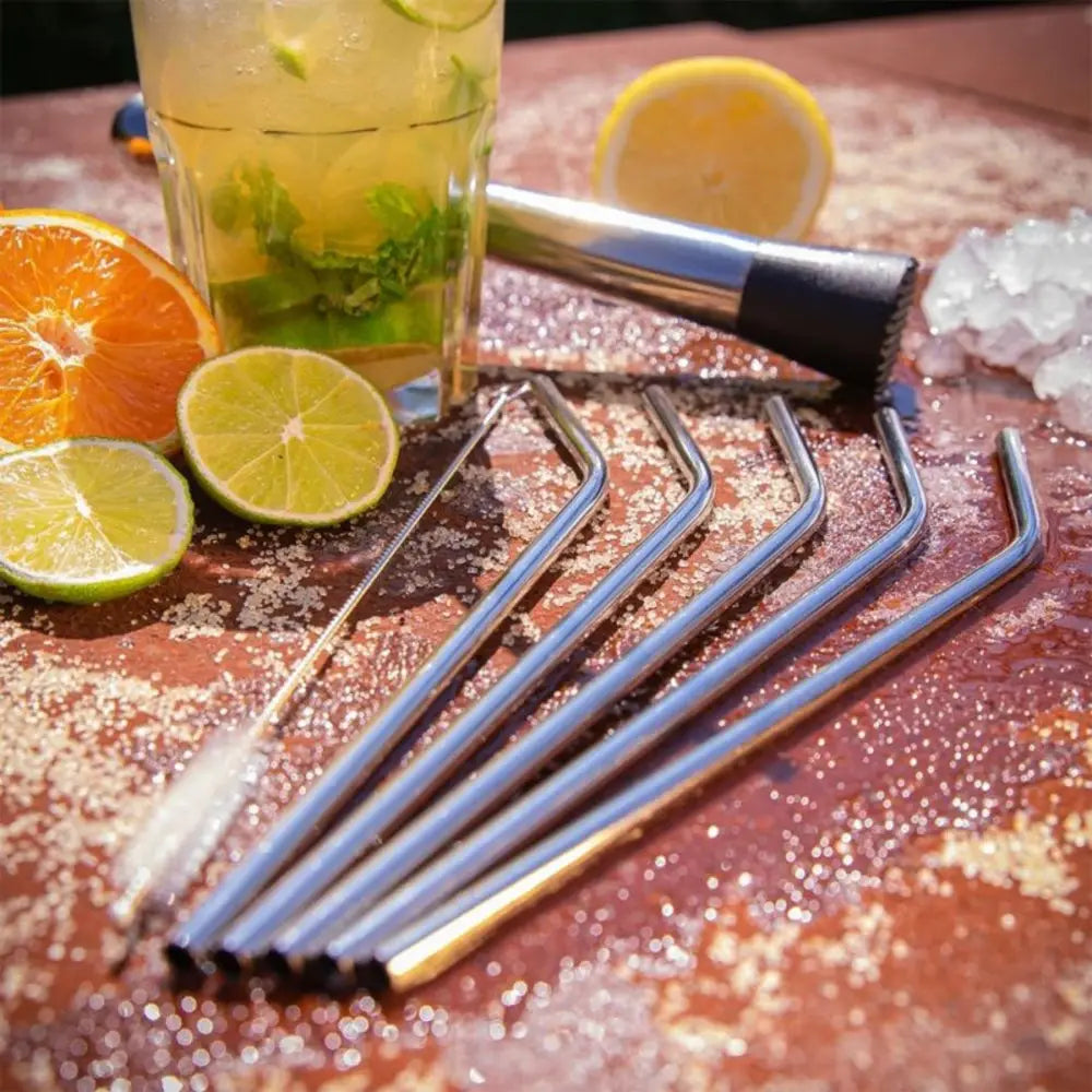 Set of metal straws and cleaning brush on a wooden surface with a cocktail and citrus fruits.