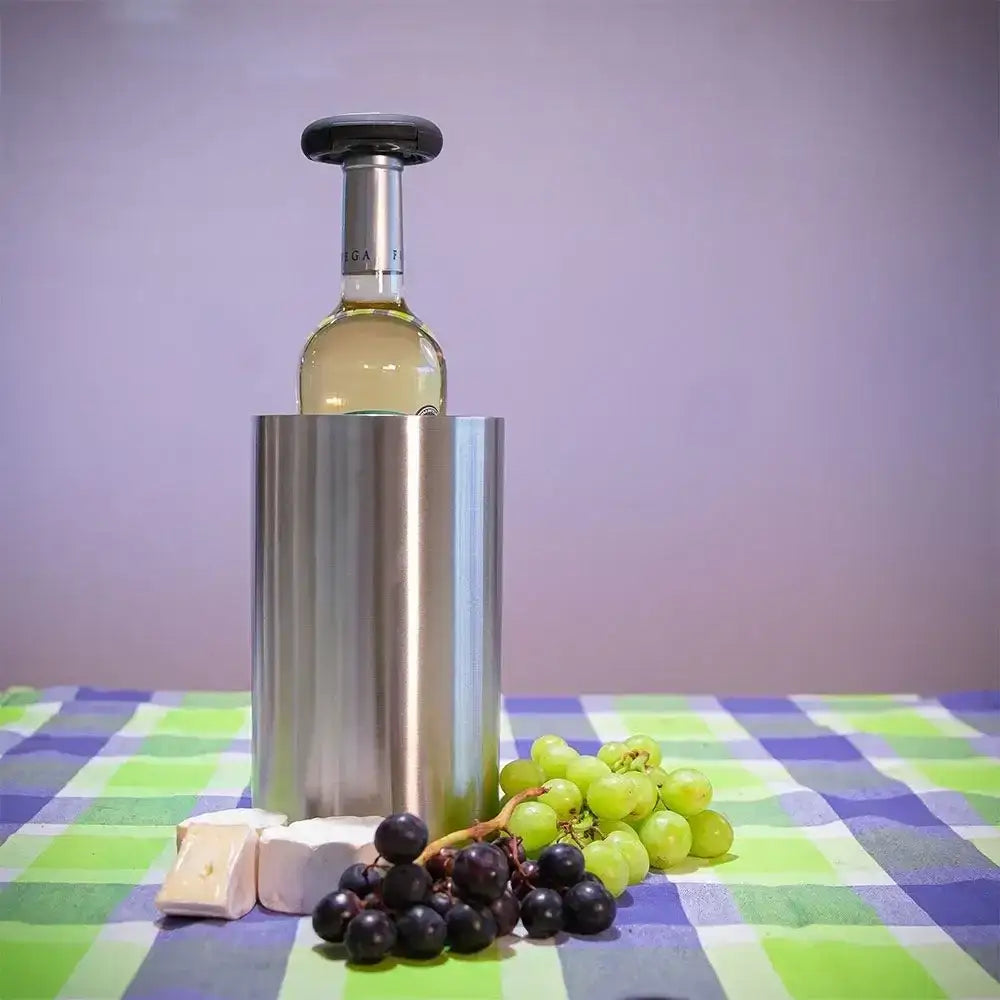 Bottle of white wine in a metal cooler with grapes and cheese on a checkered tablecloth.